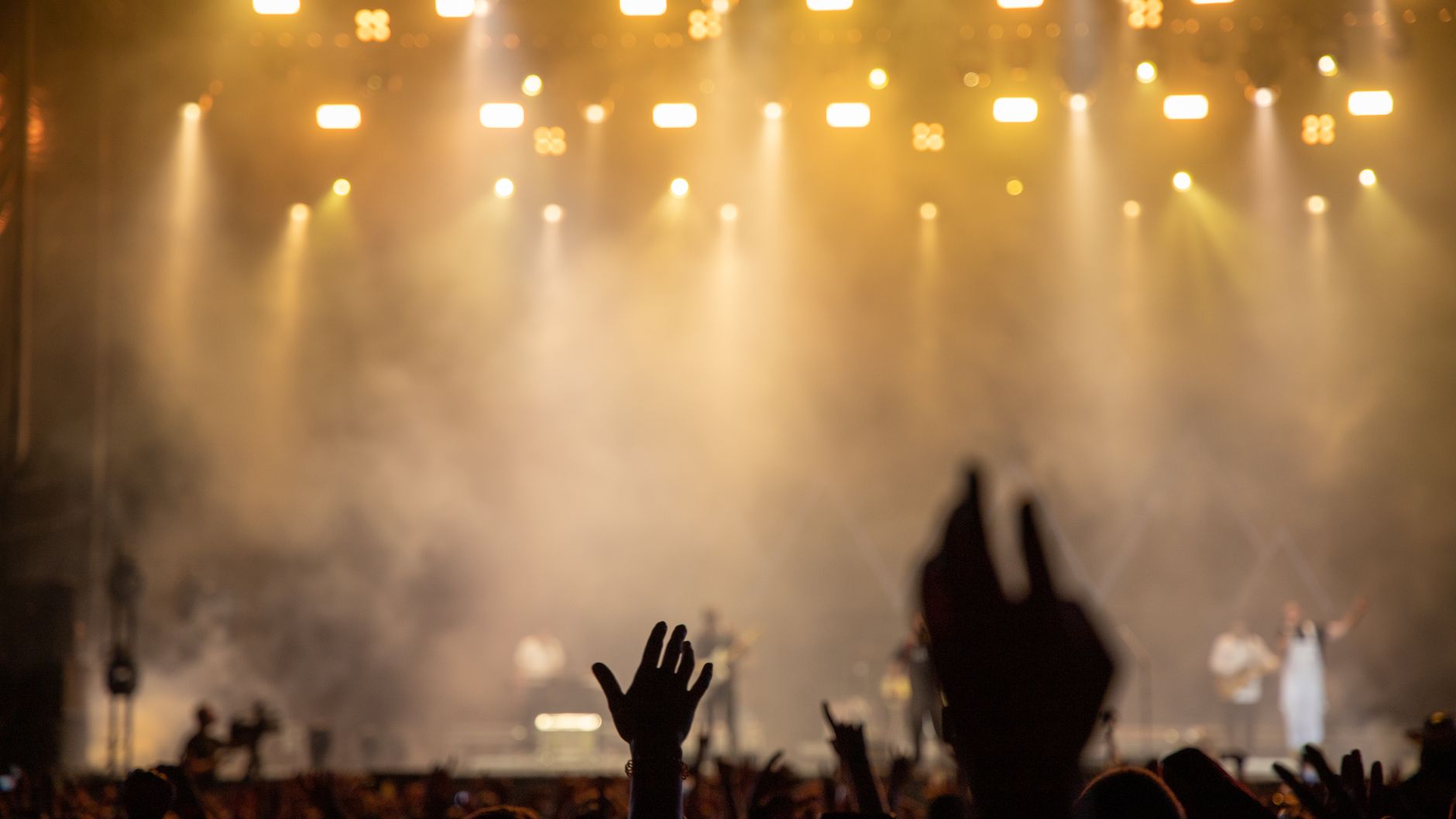 Raised hands silhouetted against warm stage lights and haze at an outdoor concert (illustrative stock photo).