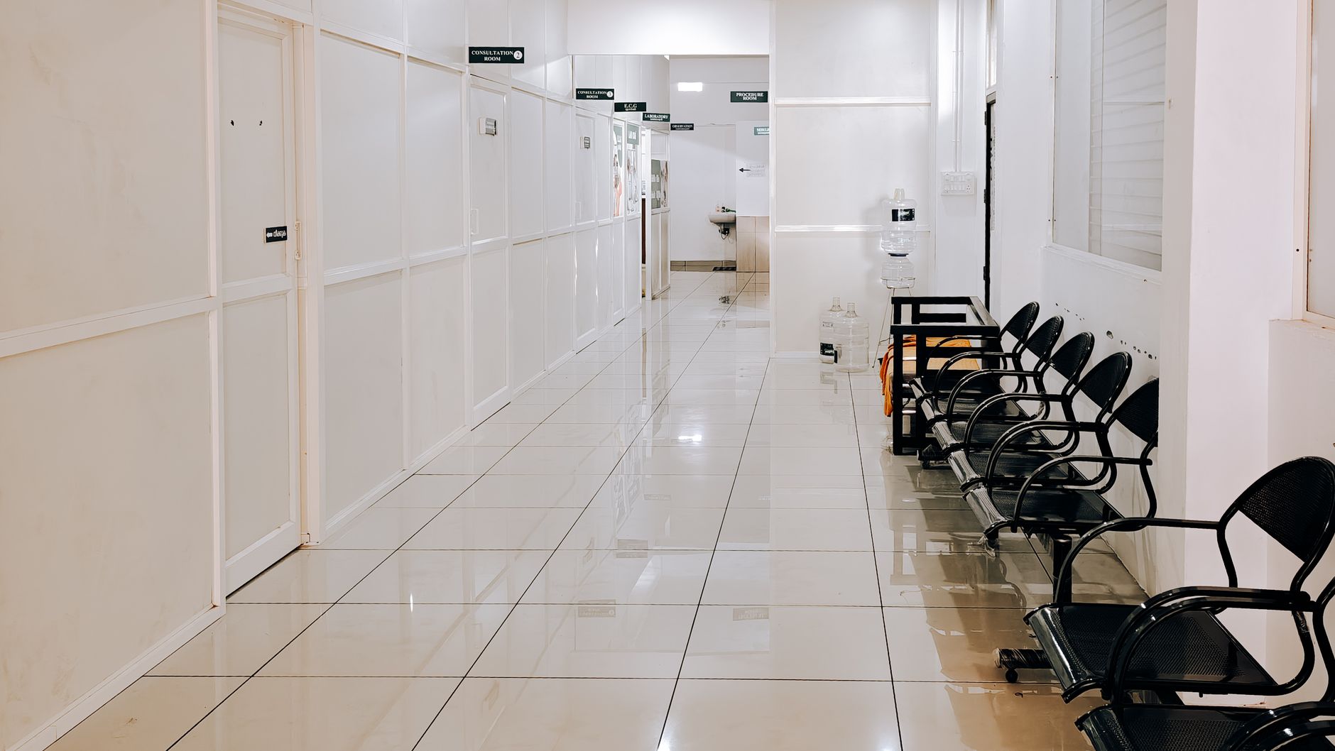 A bright, empty clinic corridor with consultation room doors on one side and a row of black waiting chairs on the other (illustrative stock photo).