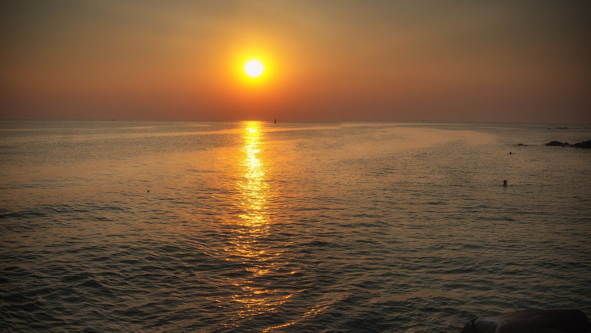 The sun low over a calm sea at dusk, with dark rocks in the foreground.