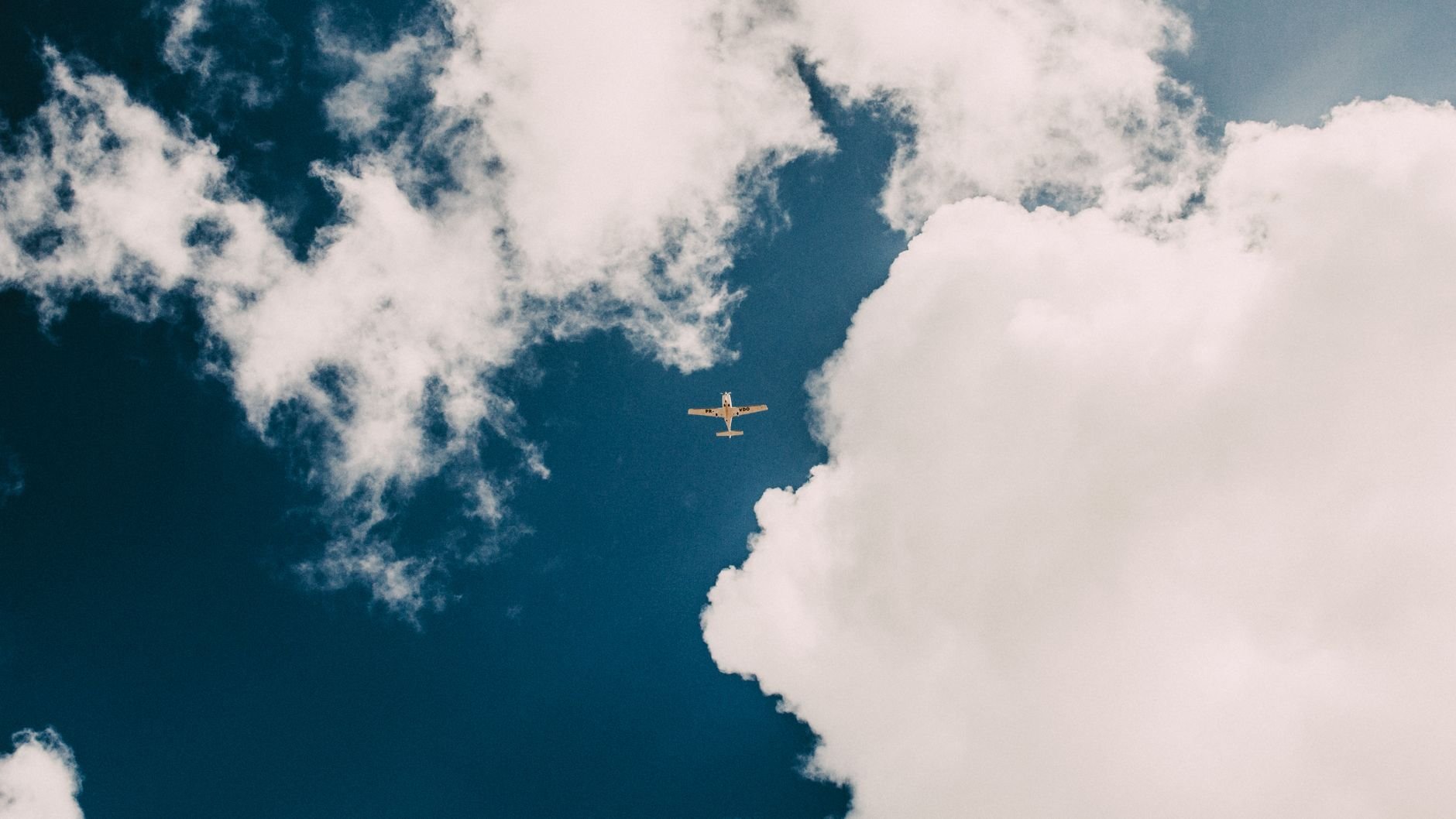 A small propeller aircraft seen from below against blue sky and large white clouds (illustrative stock photo).