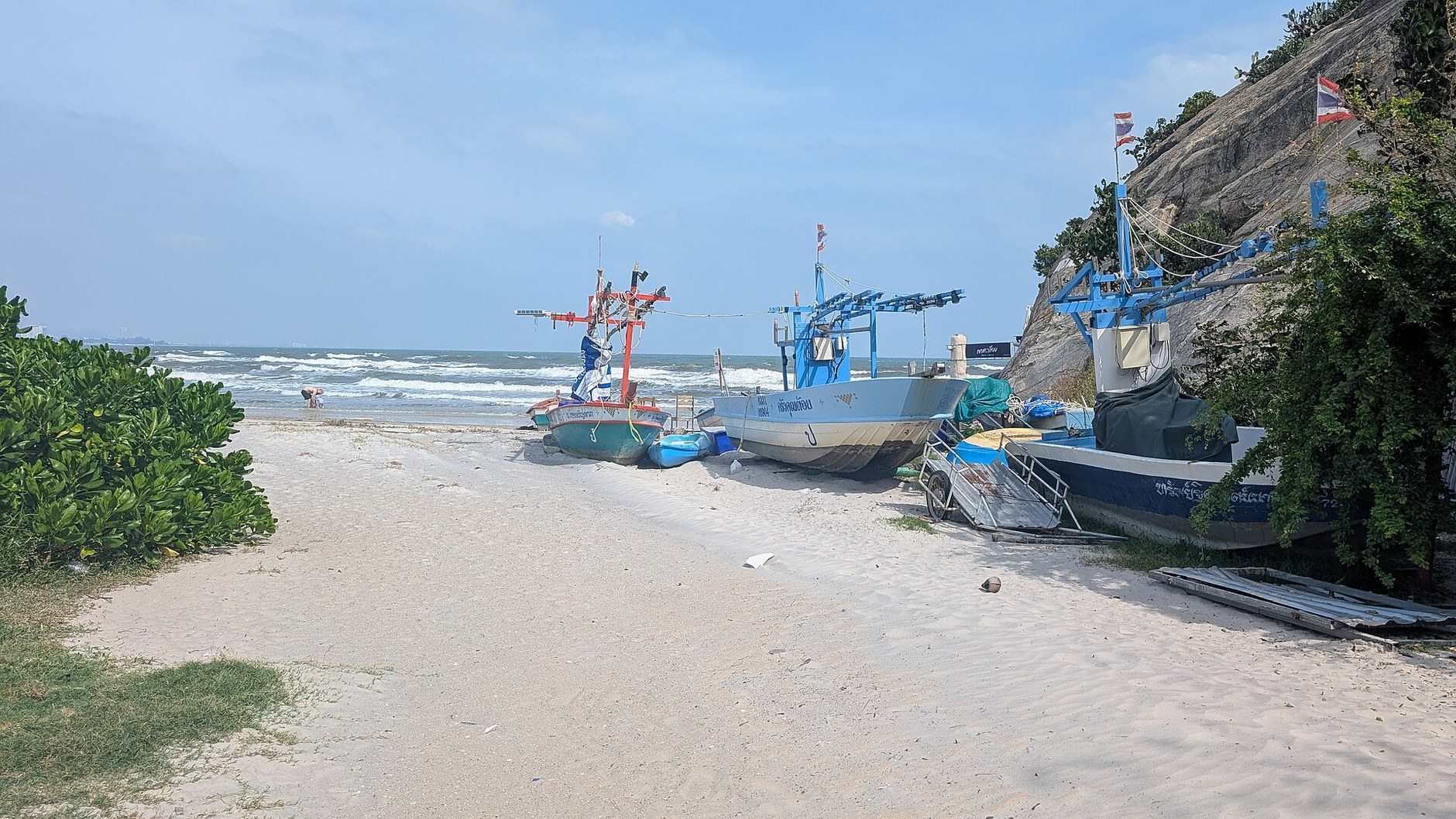 Fishing boats pulled up on a sandy Hua Hin beach beside a rocky headland, with a few ripples of surf behind.